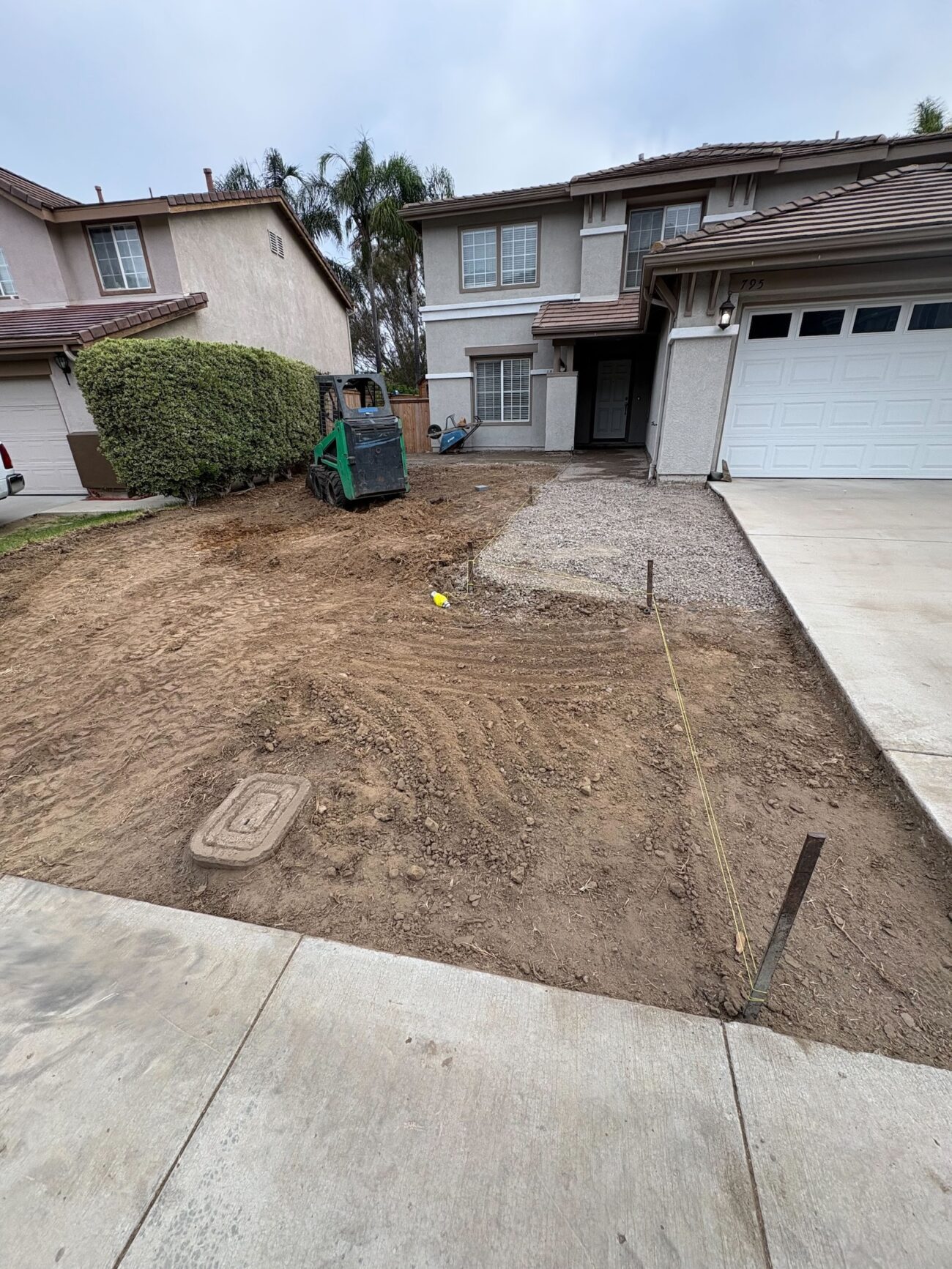 Front yard of a single-family home with existing sod removed and subgrade exposed. A small loader sits on the site; gravel has been placed adjacent to the driveway indicating base preparation for a future paver or turf installation.