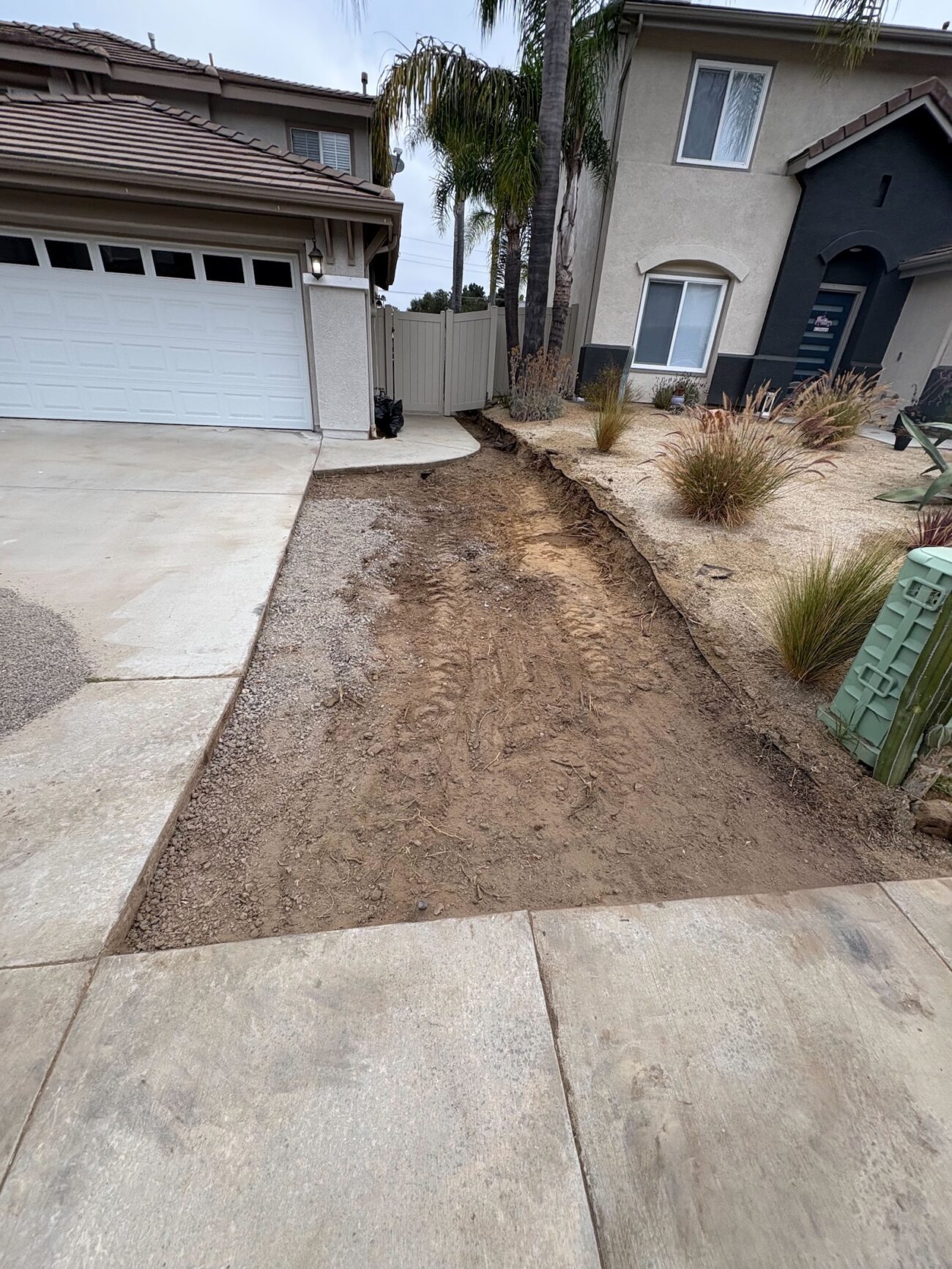 Front yard strip adjacent to the driveway has been excavated and graded in preparation for a paver walkway or driveway extension. Visible soil, track marks, and remaining planting bed indicate base preparation work underway next to the house exterior.