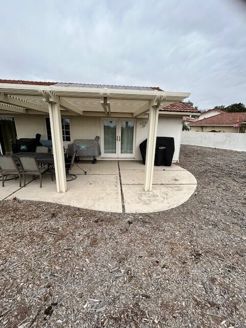 Bare concrete patio with covered pergola, patio furniture, and grills behind a stucco home before renovation.