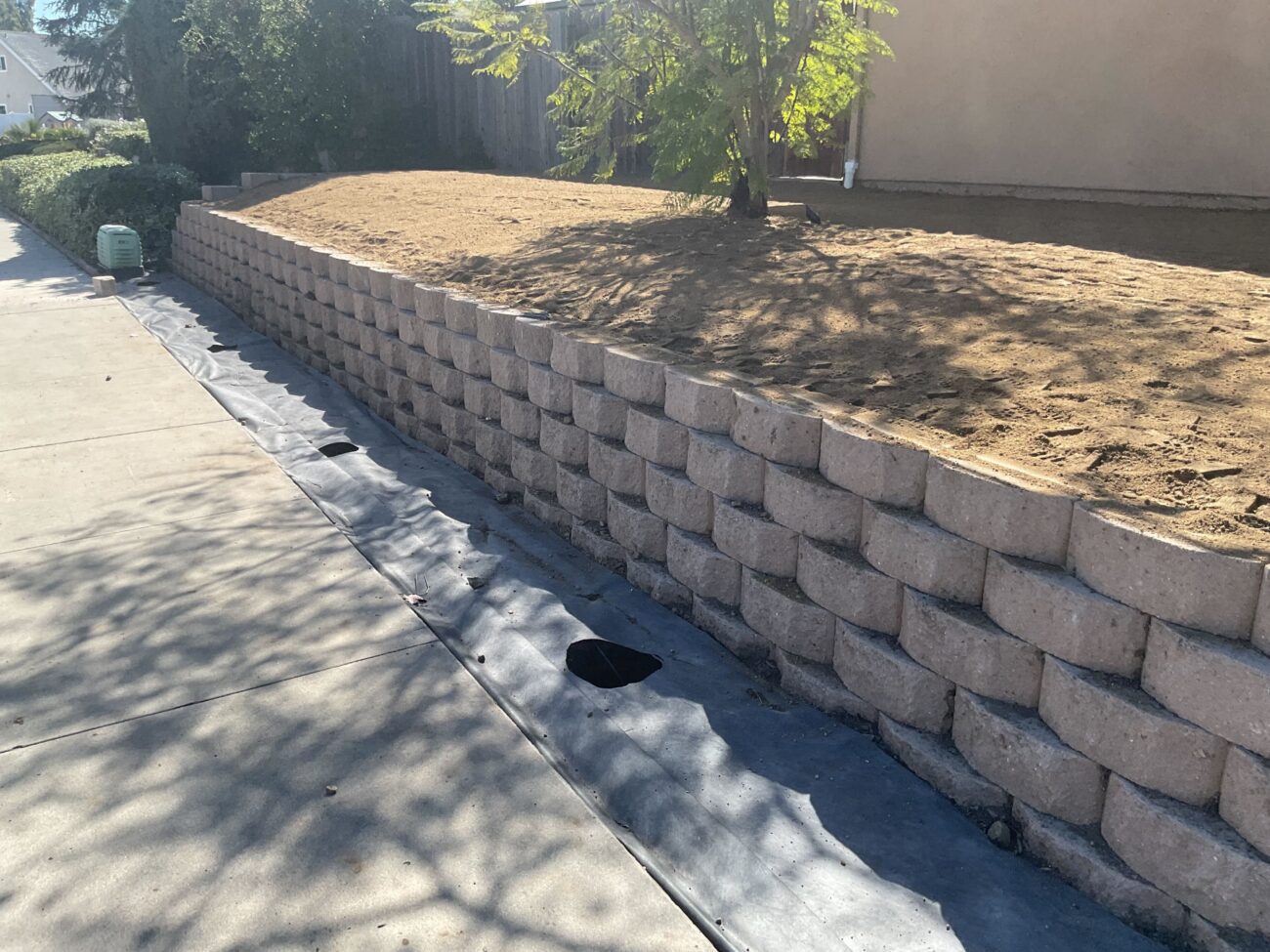 Segmental concrete block retaining wall along a residential side yard with compacted bare soil behind it. Geotextile...