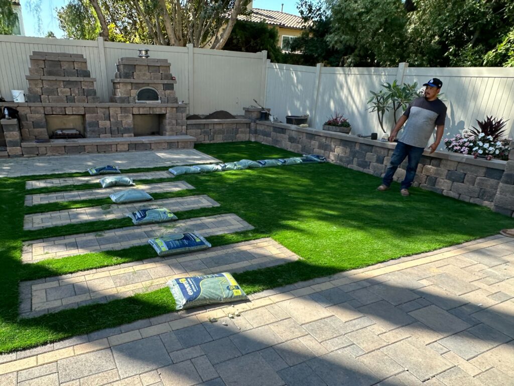 Backyard renovation showing artificial turf being installed with paver stepping stones and multiple infill bags staged on...