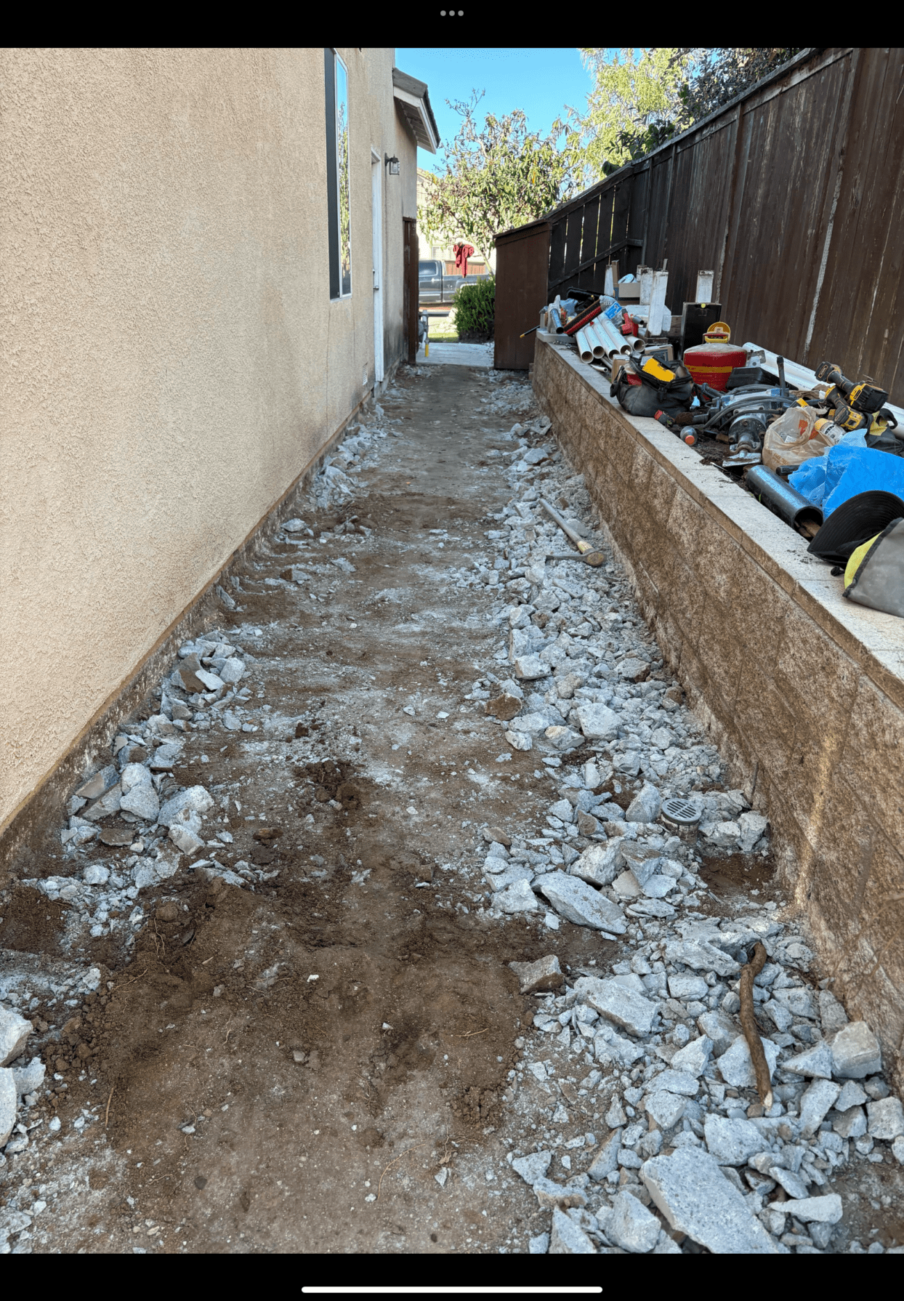 Narrow side yard walkway shows removed concrete with broken rubble and exposed subgrade running between the house and a segmental retaining wall. Tools and materials are staged on the wall coping, indicating concrete demolition in preparation for base prep and future paver installation.