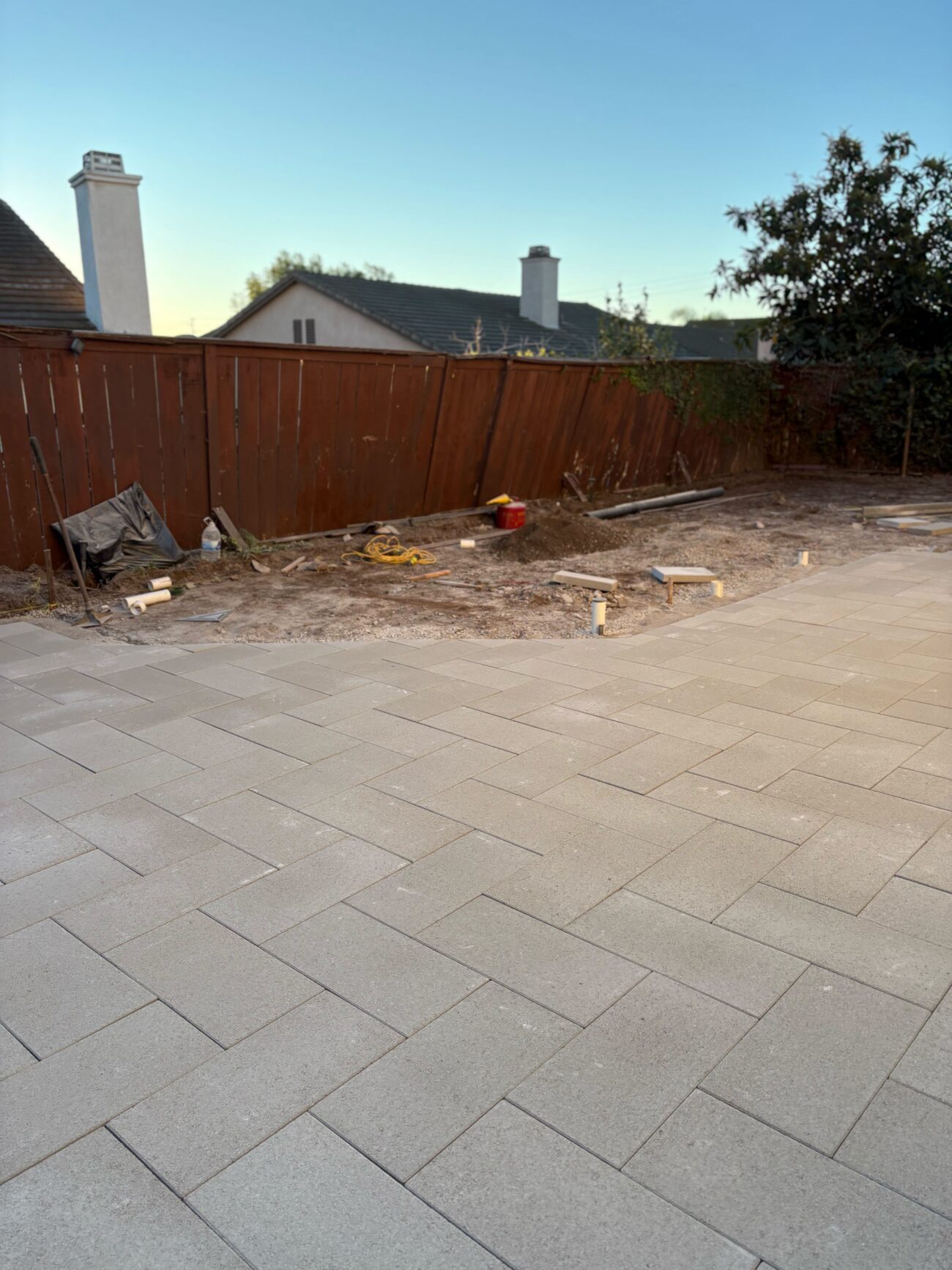 Installed paver patio in the foreground with an unfinished perimeter strip of exposed soil and base material along a...