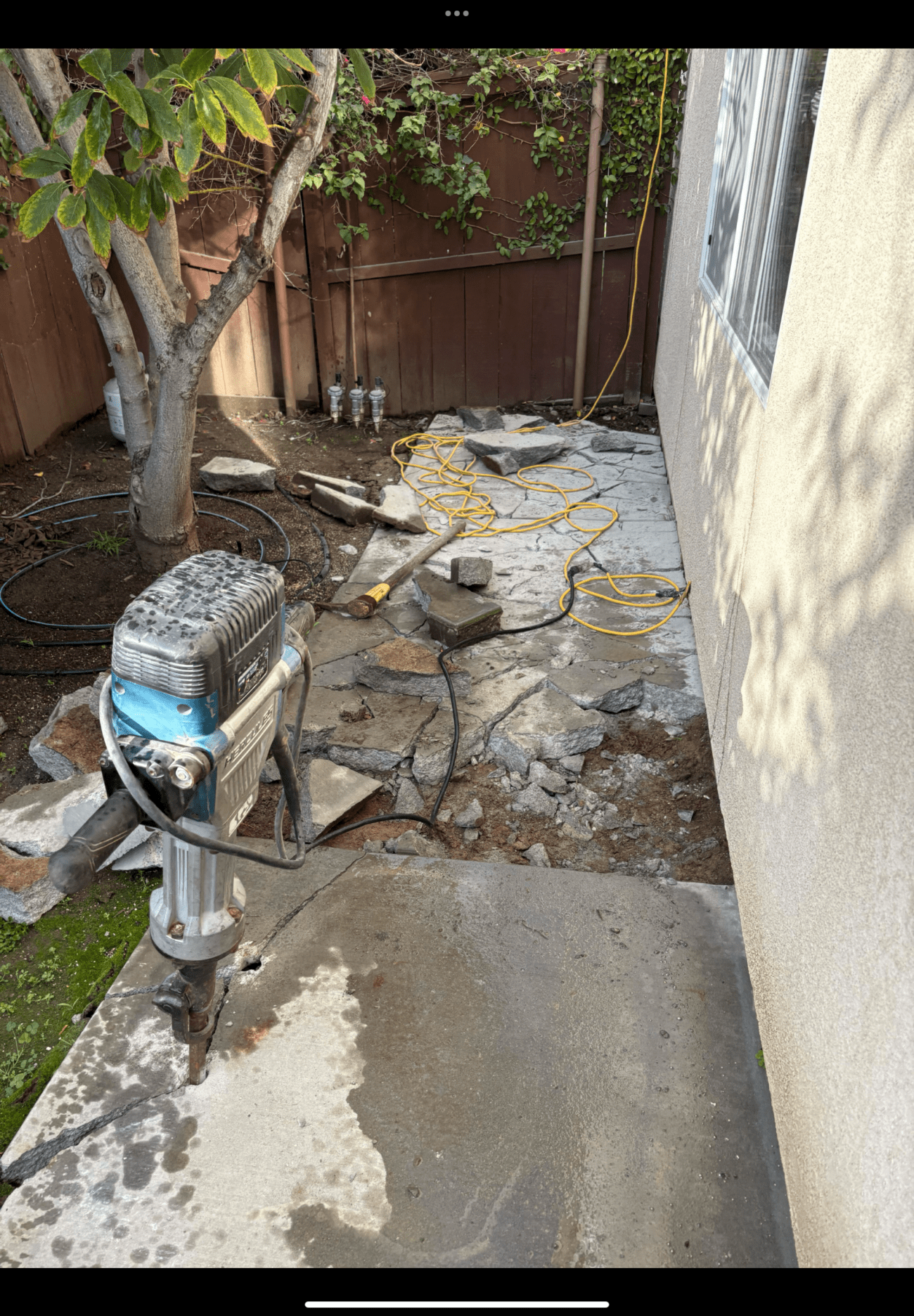 Narrow side yard undergoing concrete slab demolition. A jackhammer sits on a broken concrete walkway with scattered slab...