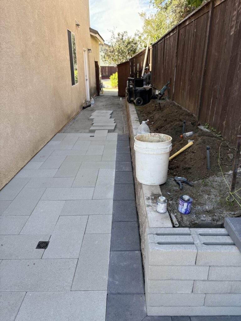 Narrow side-yard undergoing paver installation with large-format concrete pavers and a contrasting dark border being set....