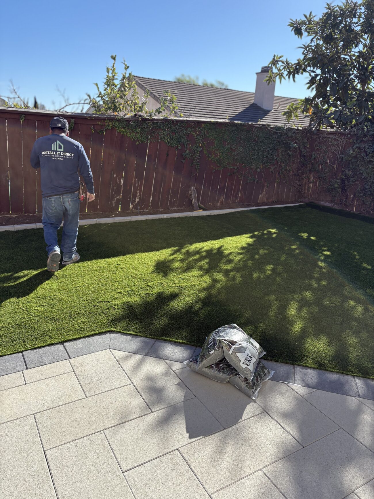 Backyard scene showing newly laid artificial turf abutting a light-colored interlocking paver patio. A crew member is on...