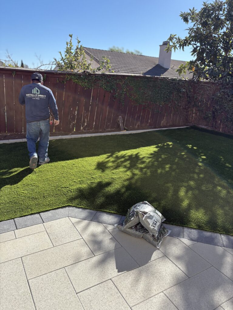 Backyard scene showing newly laid artificial turf abutting a light-colored interlocking paver patio. A crew member is on...