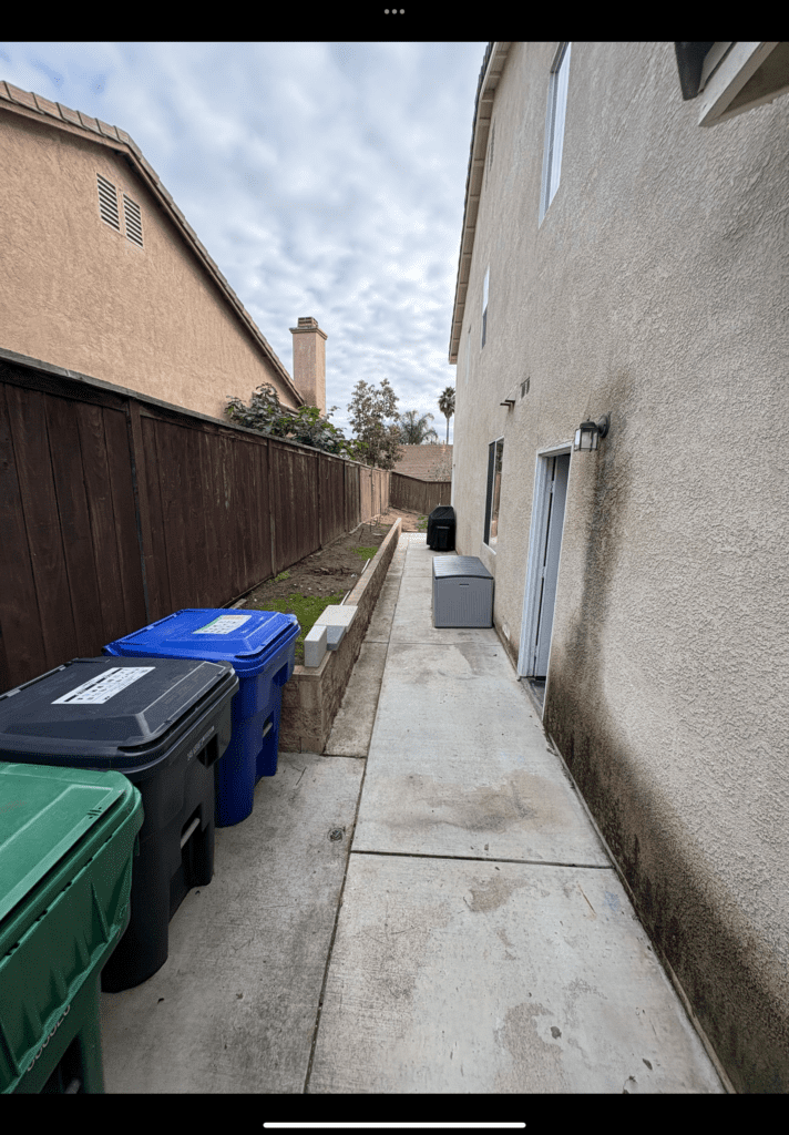 Narrow side yard with an existing concrete walkway running alongside a stucco house and a low segmental retaining the...