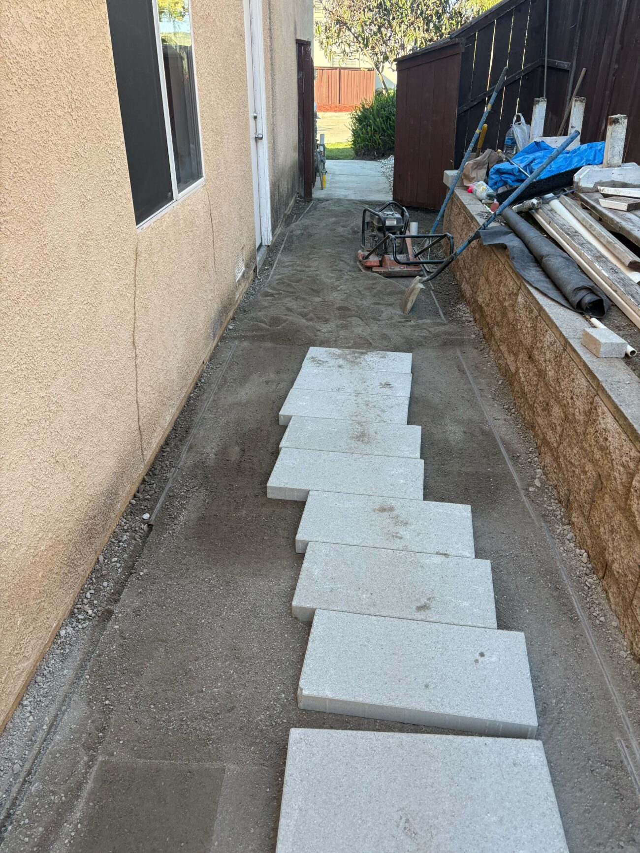 Narrow side yard walkway between the house and a retaining wall with large rectangular concrete pavers staged on a compacted aggregate base. Site shows base prep with screeded fines, stacked materials on the wall, and a plate compactor and tools at the far end ready for paver installation.