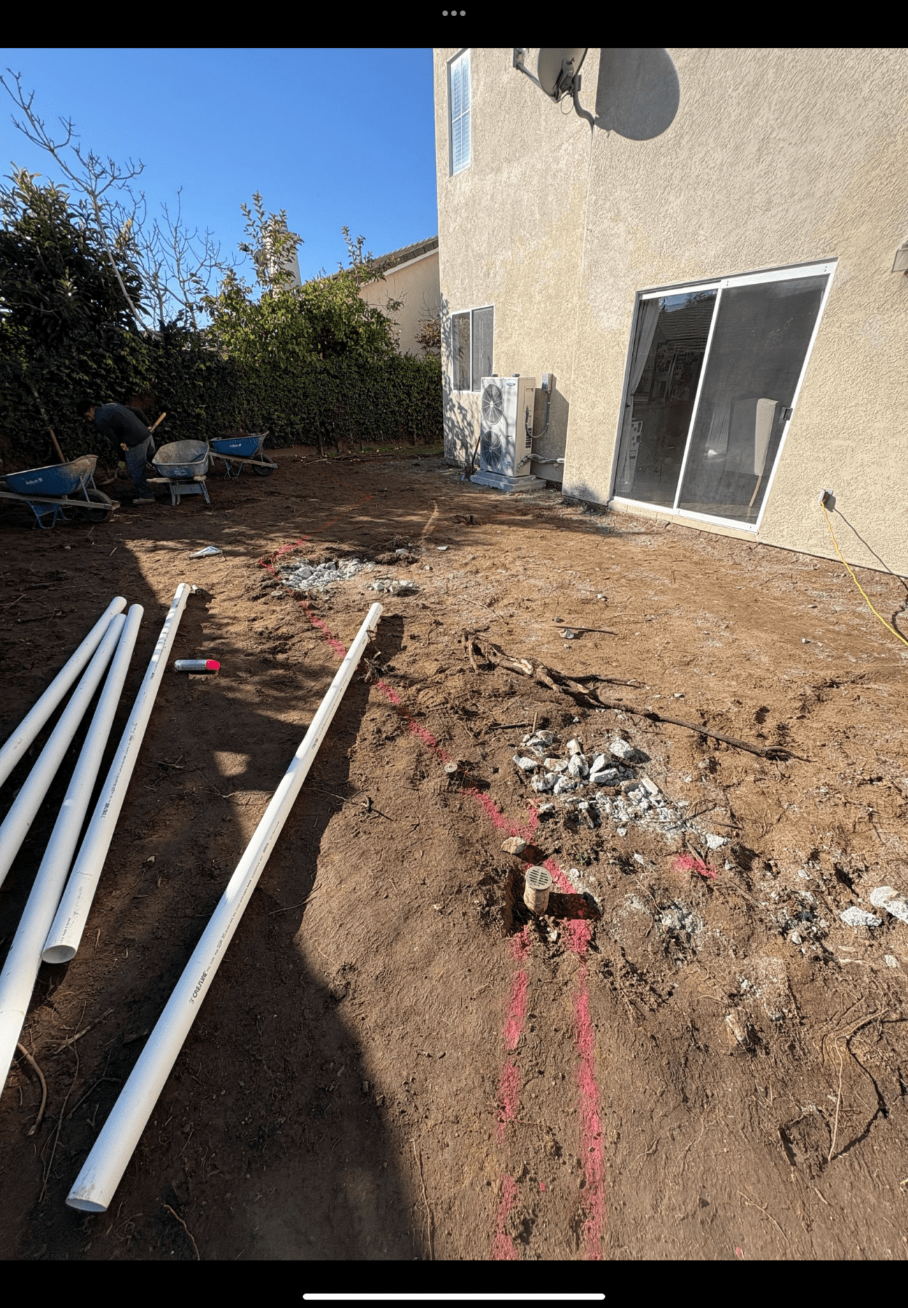Cleared residential backyard with exposed subgrade and several lengths of PVC piping staged on the soil; spray-painted...