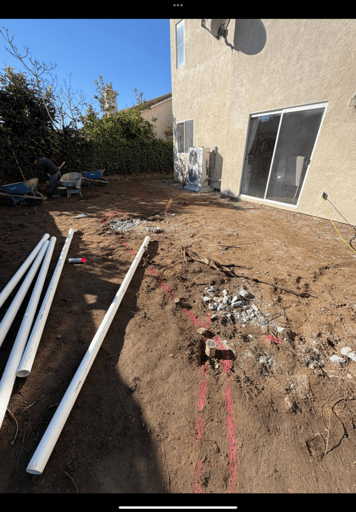 Cleared residential backyard with exposed subgrade and several lengths of PVC piping staged on the soil; spray-painted...