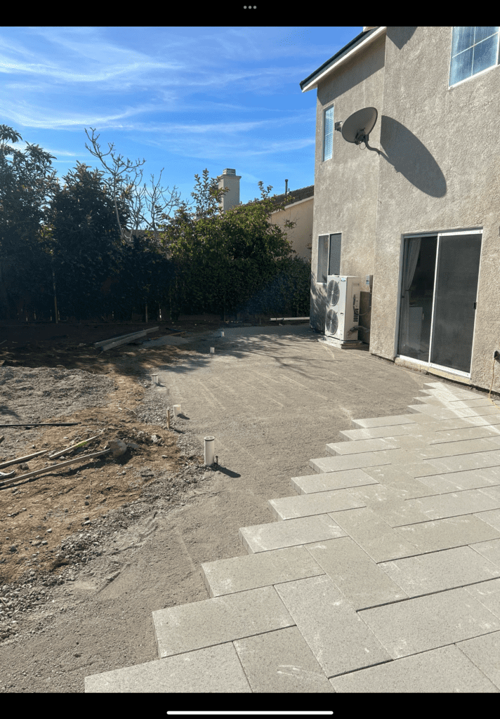 Partial installation of a residential paver patio adjacent to a house with sliding glass doors. Large rectangular...