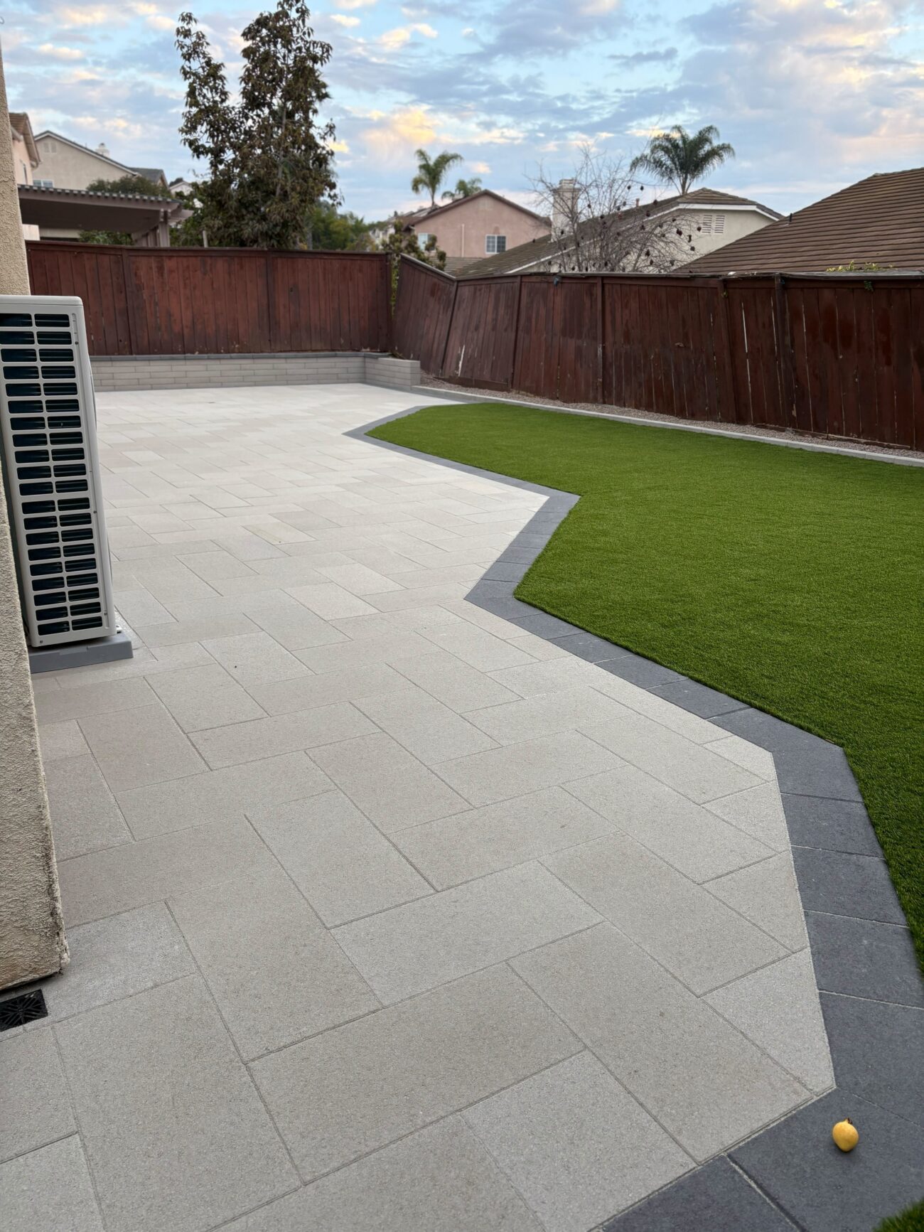 Finished residential backyard showing a light-gray interlocking paver patio with a contrasting dark border and a shaped...