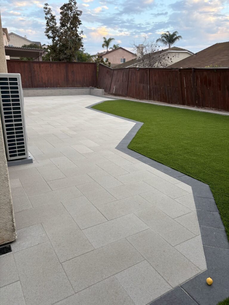 Finished residential backyard showing a light-gray interlocking paver patio with a contrasting dark border and a shaped...