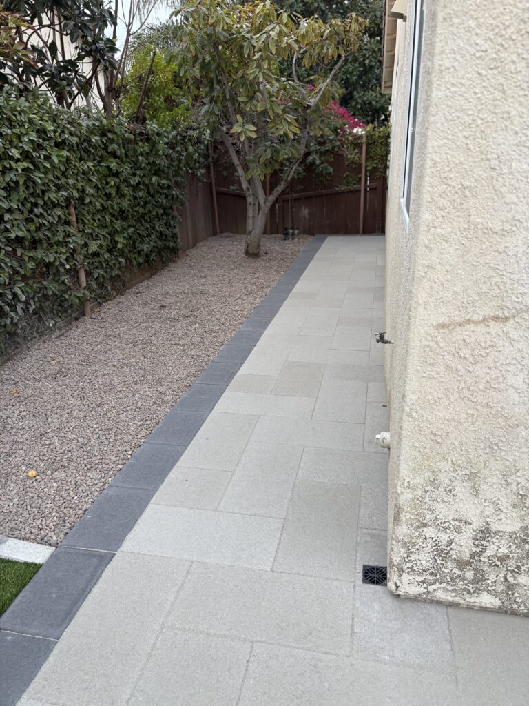 Side yard showing a completed paver walkway of light-colored square pavers with a dark border and integrated drain grate....