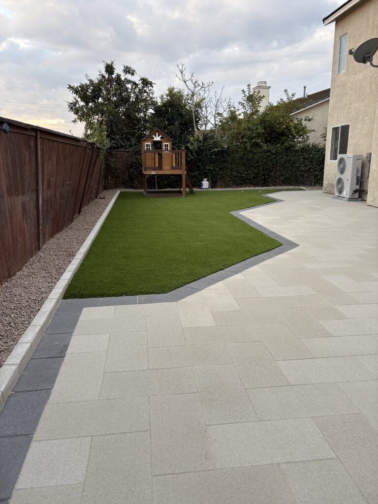 Finished backyard showing a large-format light-colored paver patio with a contrasting gray paver border and a precise...