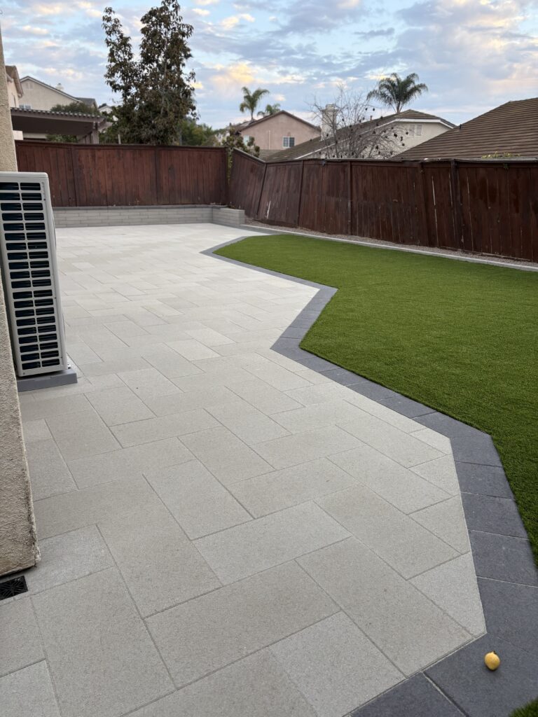 Finished residential backyard showing a large light-gray paver patio with a dark contrasting border and an adjacent...