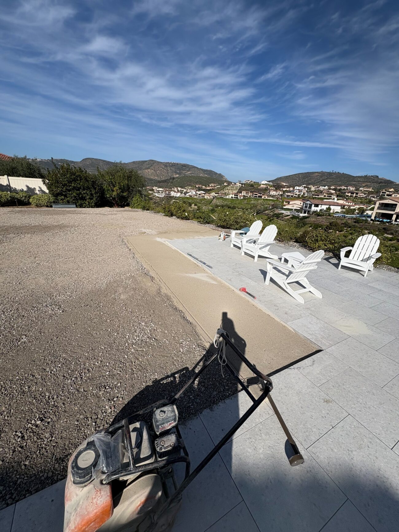 Residential paver patio in active base prep showing installed light-gray stone pavers with Adirondack chairs on the...