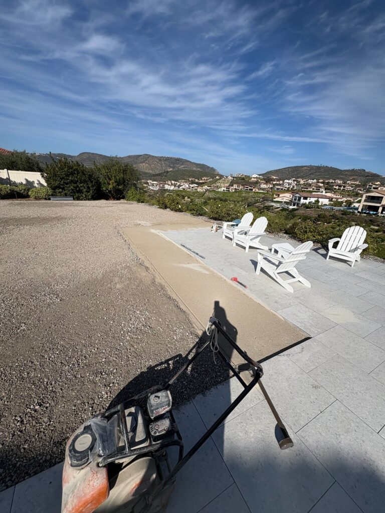 Residential paver patio in active base prep showing installed light-gray stone pavers with Adirondack chairs on the...