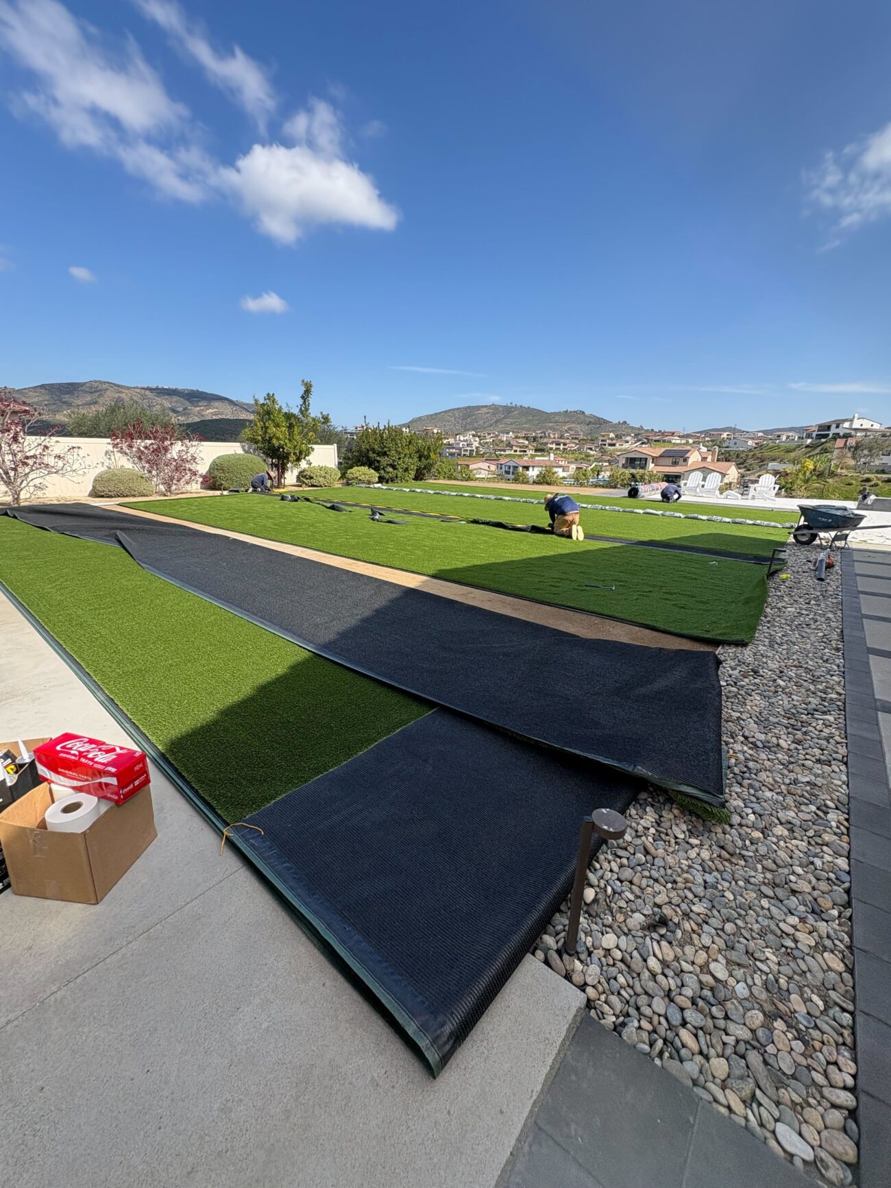 Residential backyard artificial turf installation with multiple turf rolls unrolled over the prepared base and a crew...