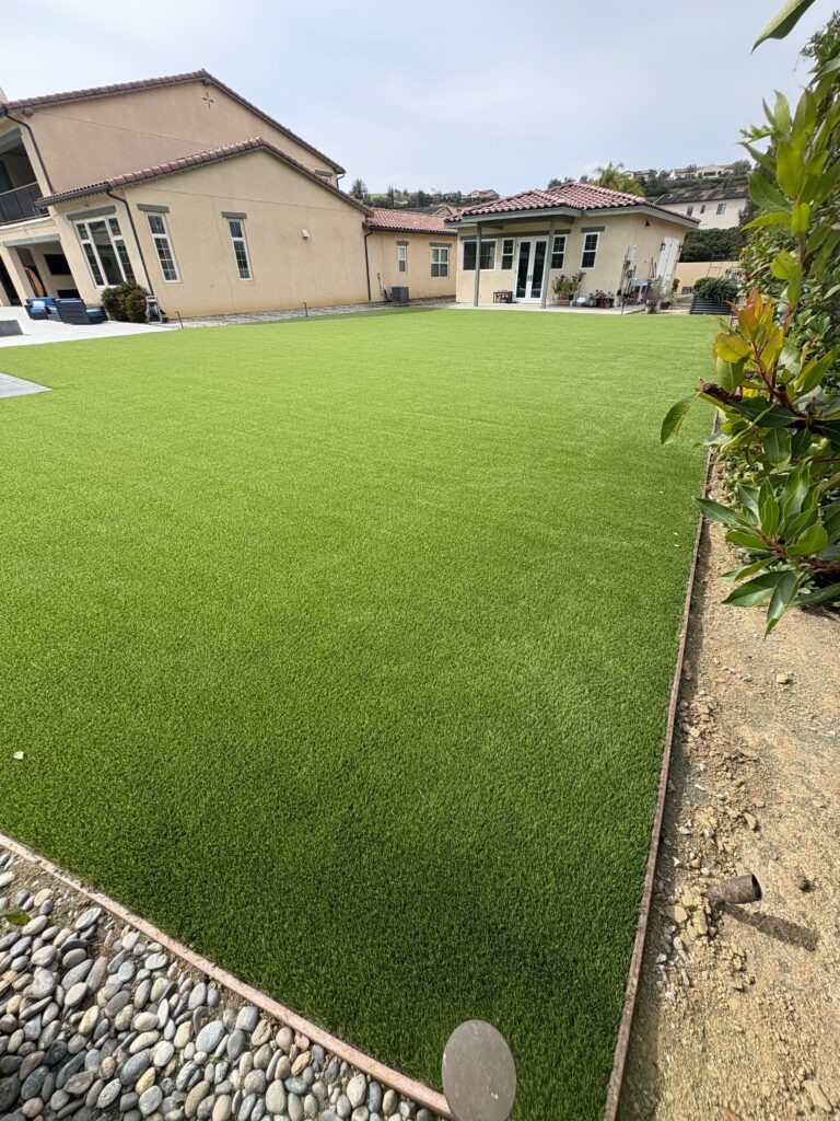Residential backyard showing a large area of freshly installed artificial turf with a visible edge restraint and adjacent...