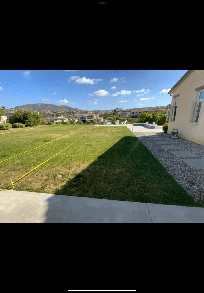 Residential backyard with an existing concrete patio edge and rock mulch along the house. Site layout strings/tape lines...