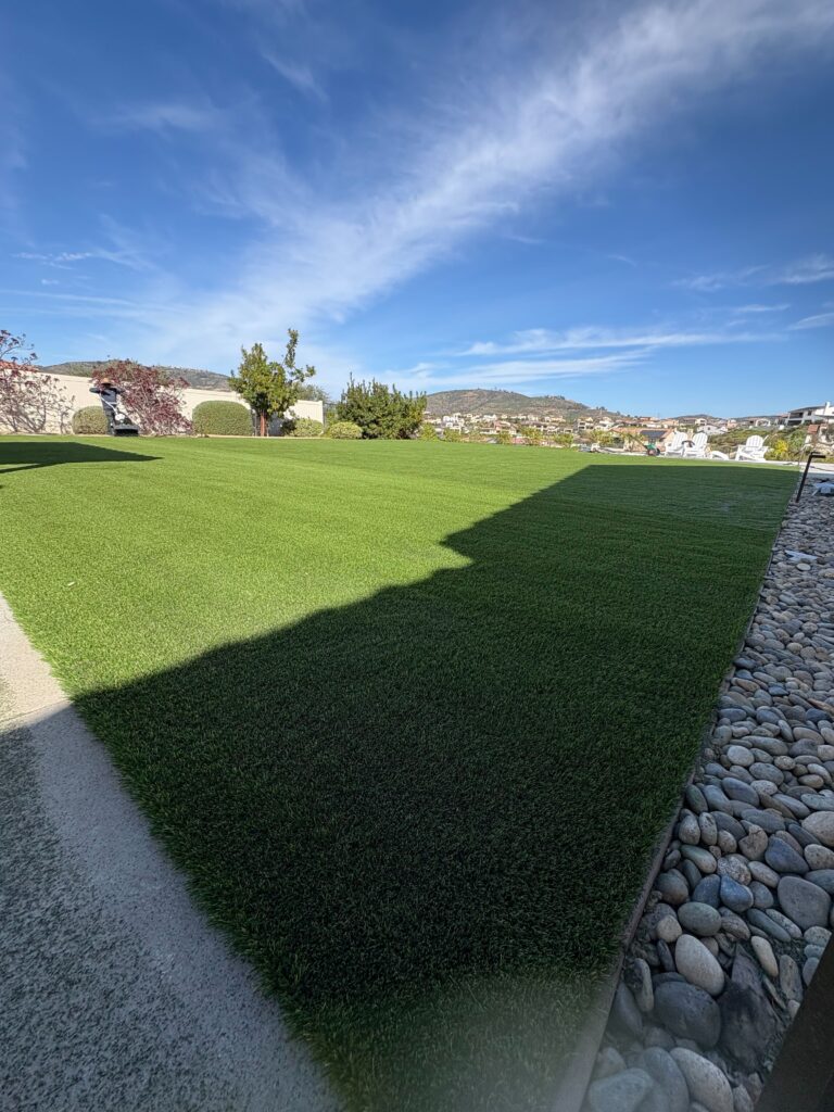 Residential backyard showing a newly installed artificial turf lawn with a neat edge against a river rock border and...
