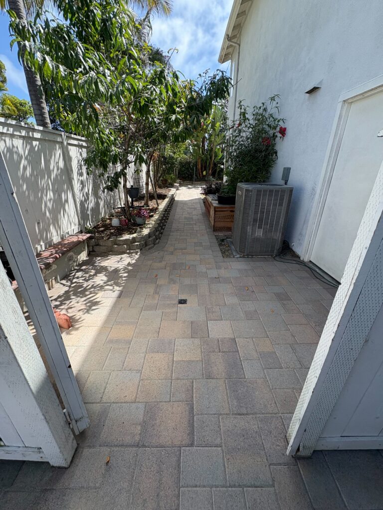 Narrow side yard with newly installed interlocking pavers forming a walkway/patio adjacent to the house. A low segmental...