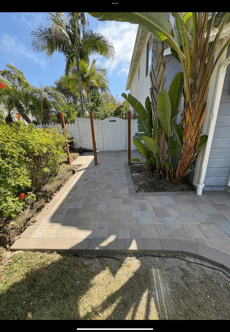 Interlocking paver walkway/patio installed along the side of a house, terminating at a white gate. Planting bed with palm...