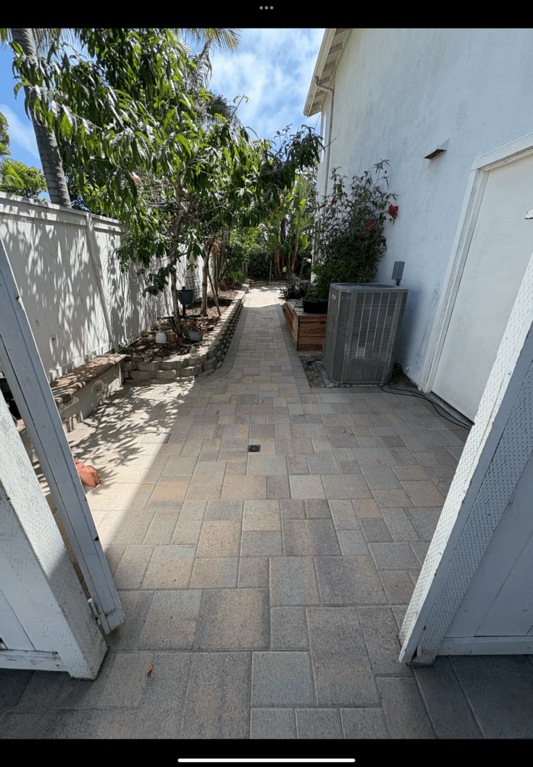 Completed interlocking concrete paver walkway installed between the house and a vinyl fence, showing a modular/patterned...