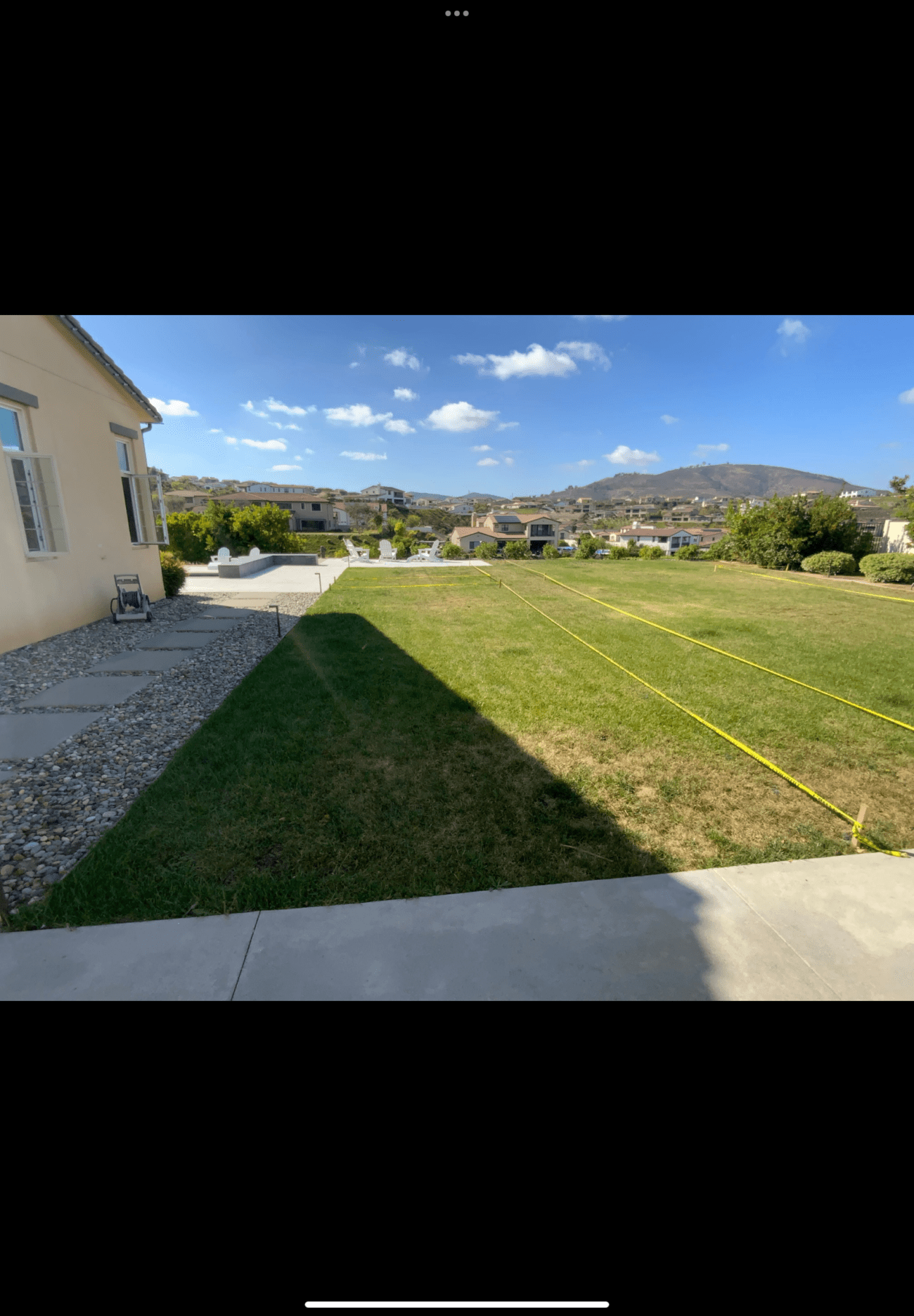 Residential backyard with an existing concrete patio edge and rock mulch along the house. Site layout strings/tape lines are stretched across the turf indicating pre-construction measurements and layout for upcoming work.
