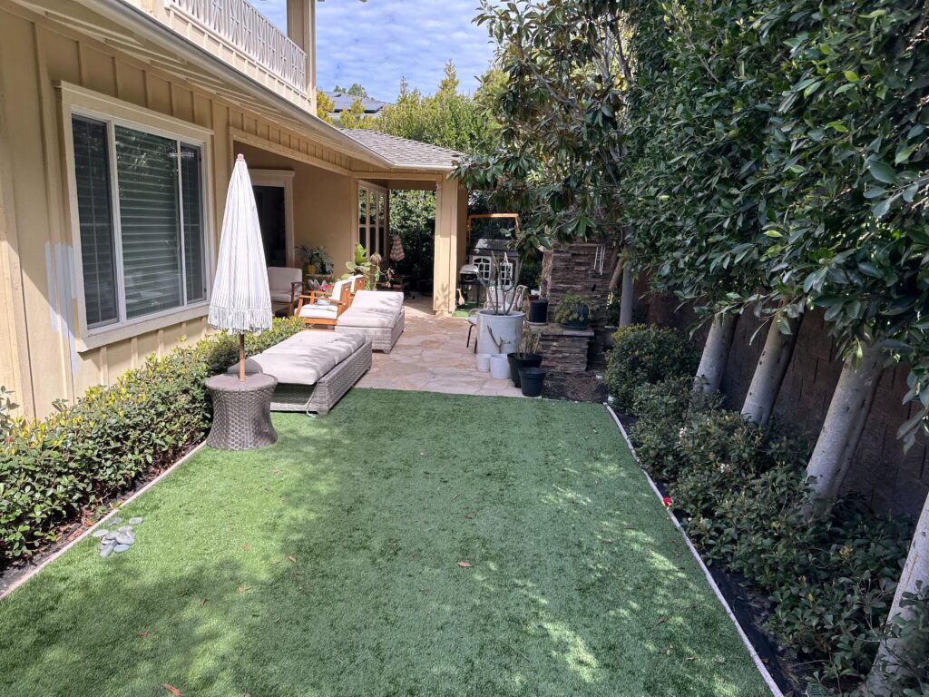 Narrow residential side yard showing newly installed artificial turf bordered by metal edging leading to a...