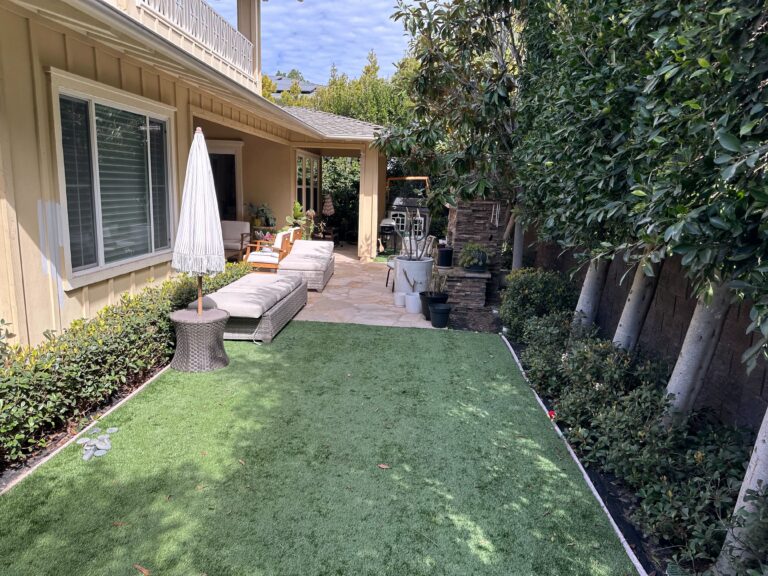 Narrow residential side yard showing newly installed artificial turf bordered by metal edging leading to a...