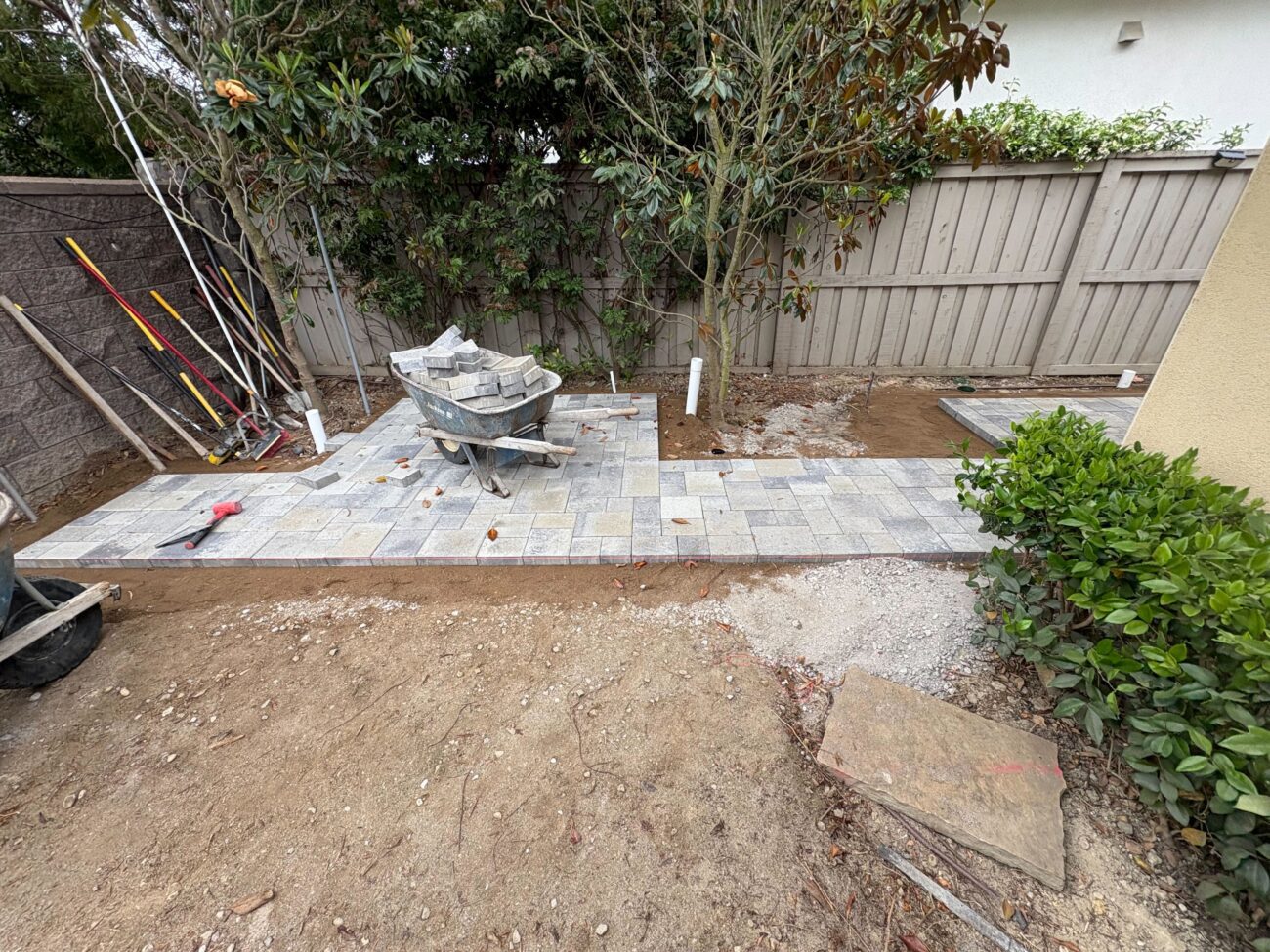 Partial installation of interlocking pavers forming a narrow patio/walkway along a house exterior. Compacted gravel base...