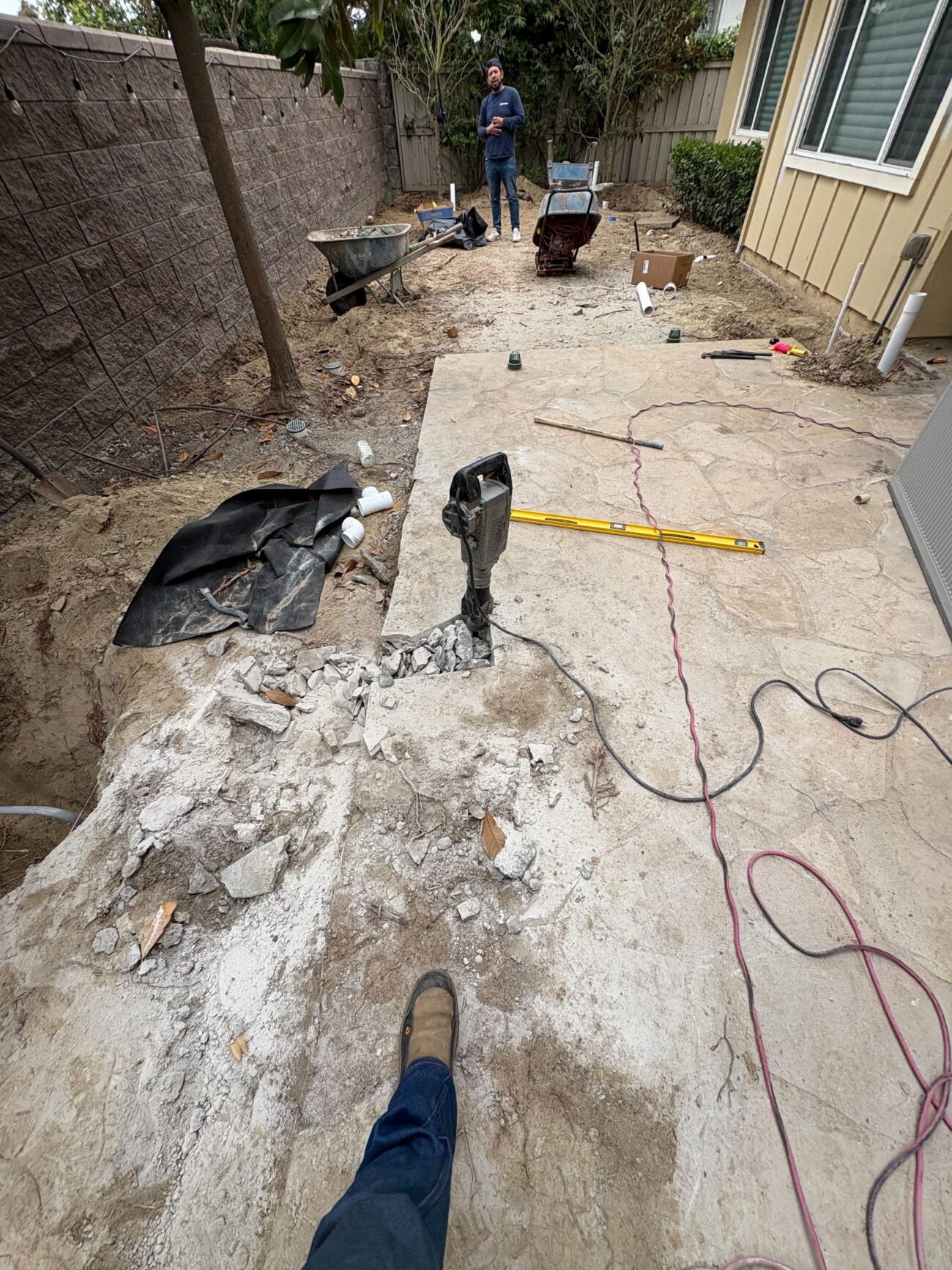 Crew is demolishing an existing concrete patio slab in a narrow backyard using a jackhammer; broken concrete and rubble...