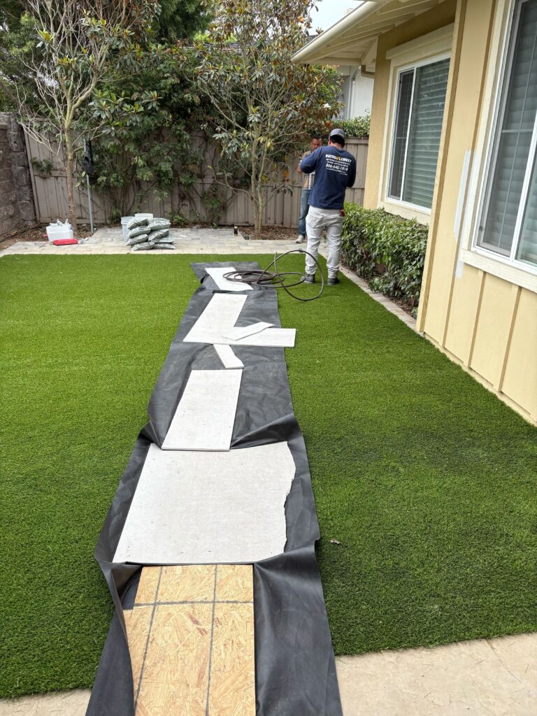 Backyard artificial turf installation in progress with turf rolls unrolled over landscape fabric and seams being...