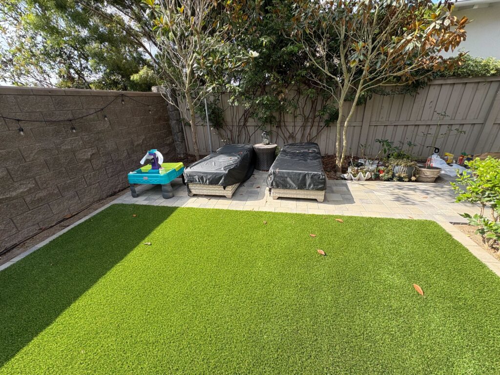 Backyard shows newly installed artificial turf in the foreground with a paved patio and segmental retaining wall along...