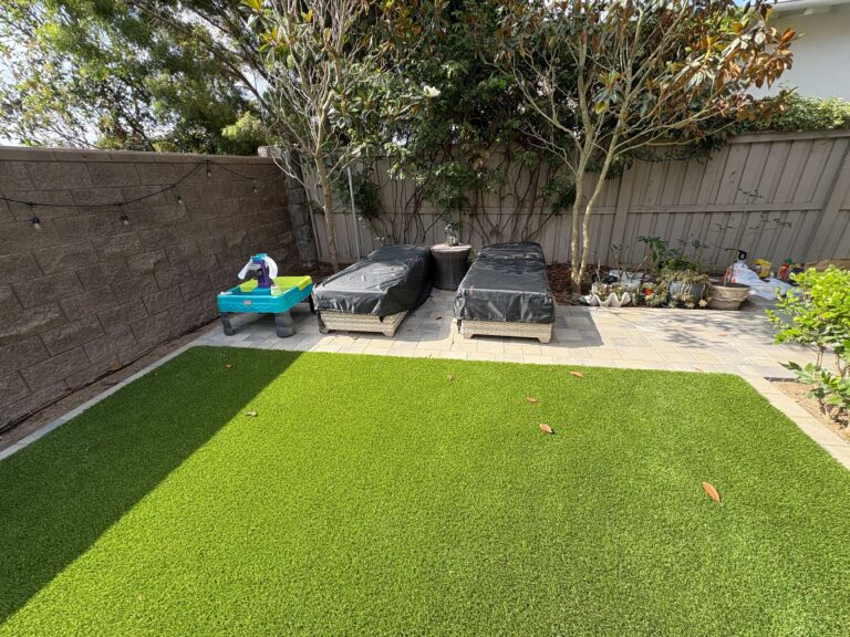 Backyard shows newly installed artificial turf in the foreground with a paved patio and segmental retaining wall along...