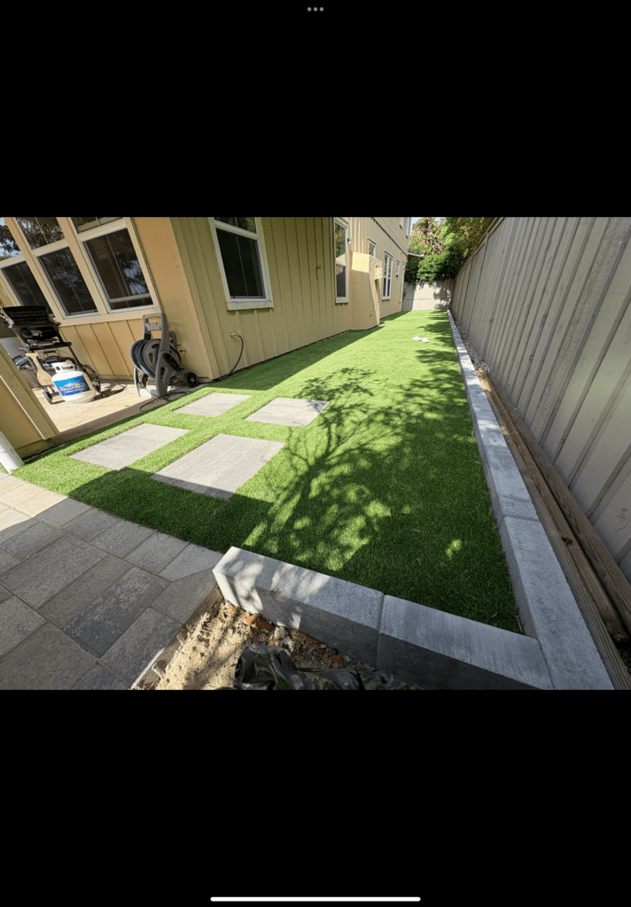 Narrow residential side yard showing a completed artificial turf installation with rectangular concrete paver stepping...