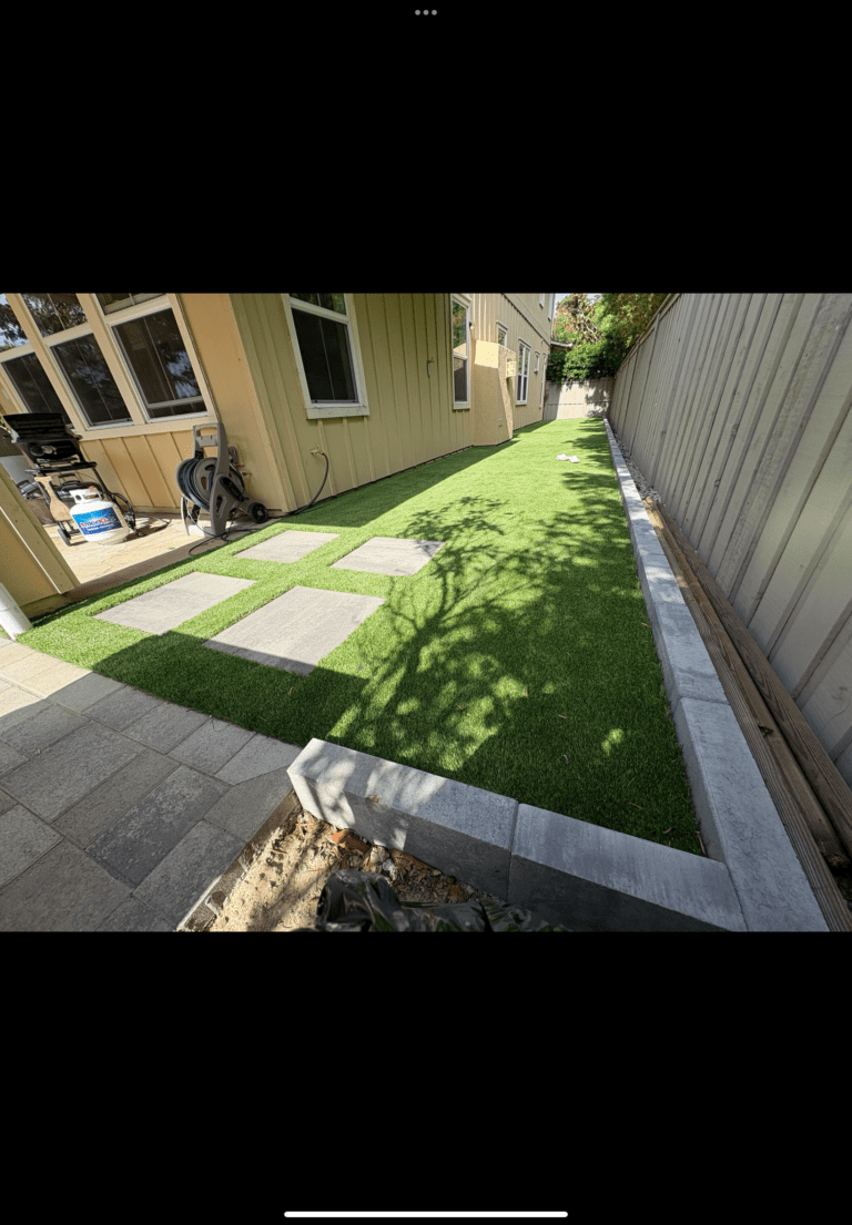 Narrow residential side yard showing a completed artificial turf installation with rectangular concrete paver stepping...
