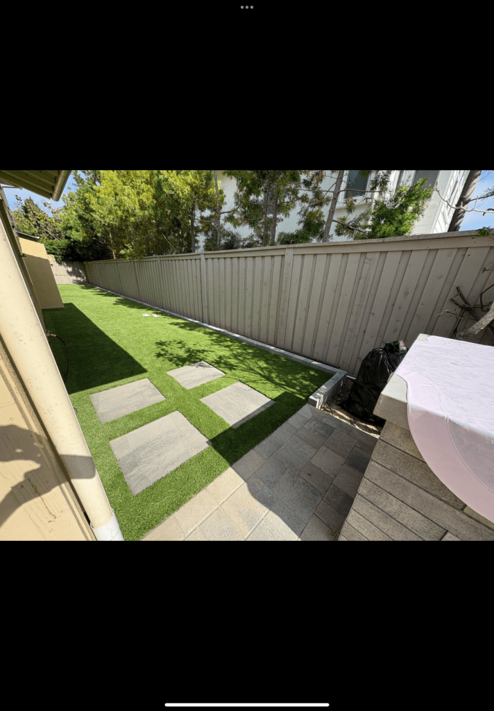 Narrow side yard shows completed artificial turf with large square paver stepping pads running from a small interlocking...