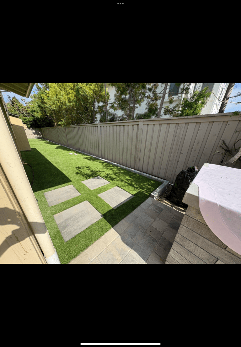 Narrow side yard shows completed artificial turf with large square paver stepping pads running from a small interlocking...
