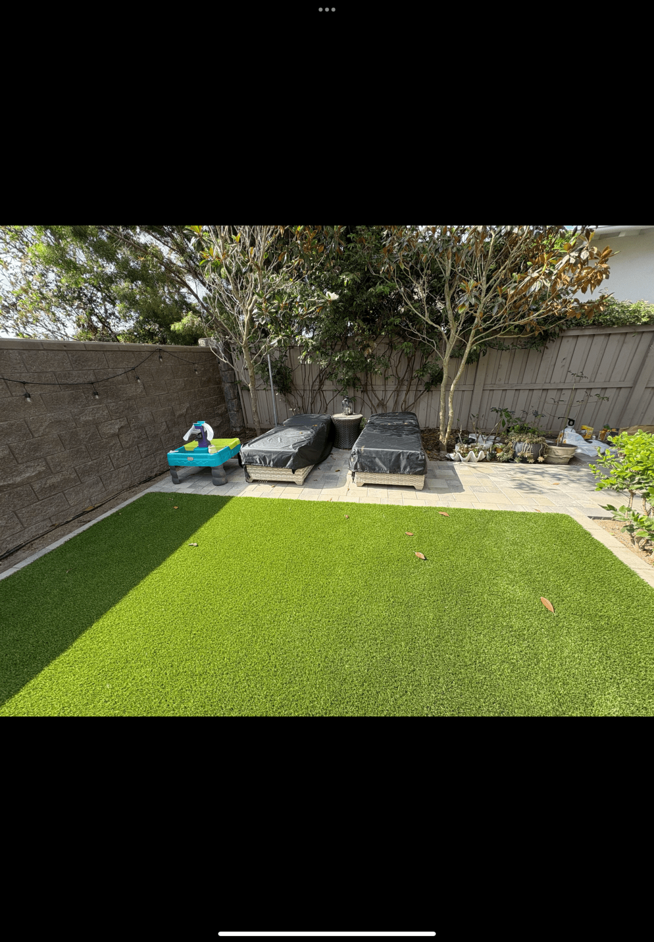 Newly installed artificial turf in a residential backyard adjacent to a paved patio/walkway and planted beds. A segmental...