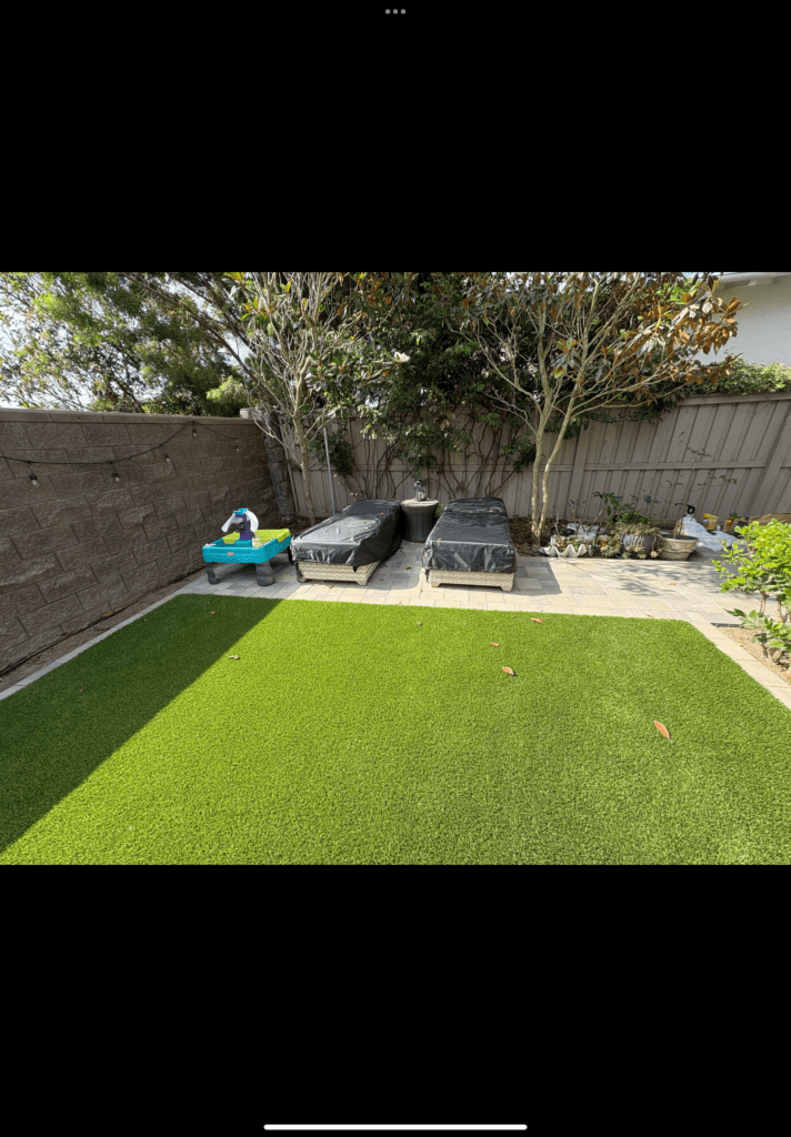 Newly installed artificial turf in a residential backyard adjacent to a paved patio/walkway and planted beds. A segmental...
