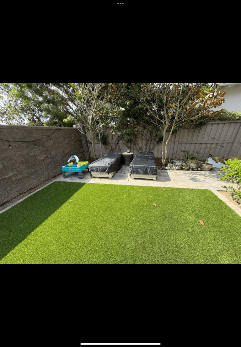 Newly installed artificial turf in a residential backyard adjacent to a paved patio/walkway and planted beds. A segmental...