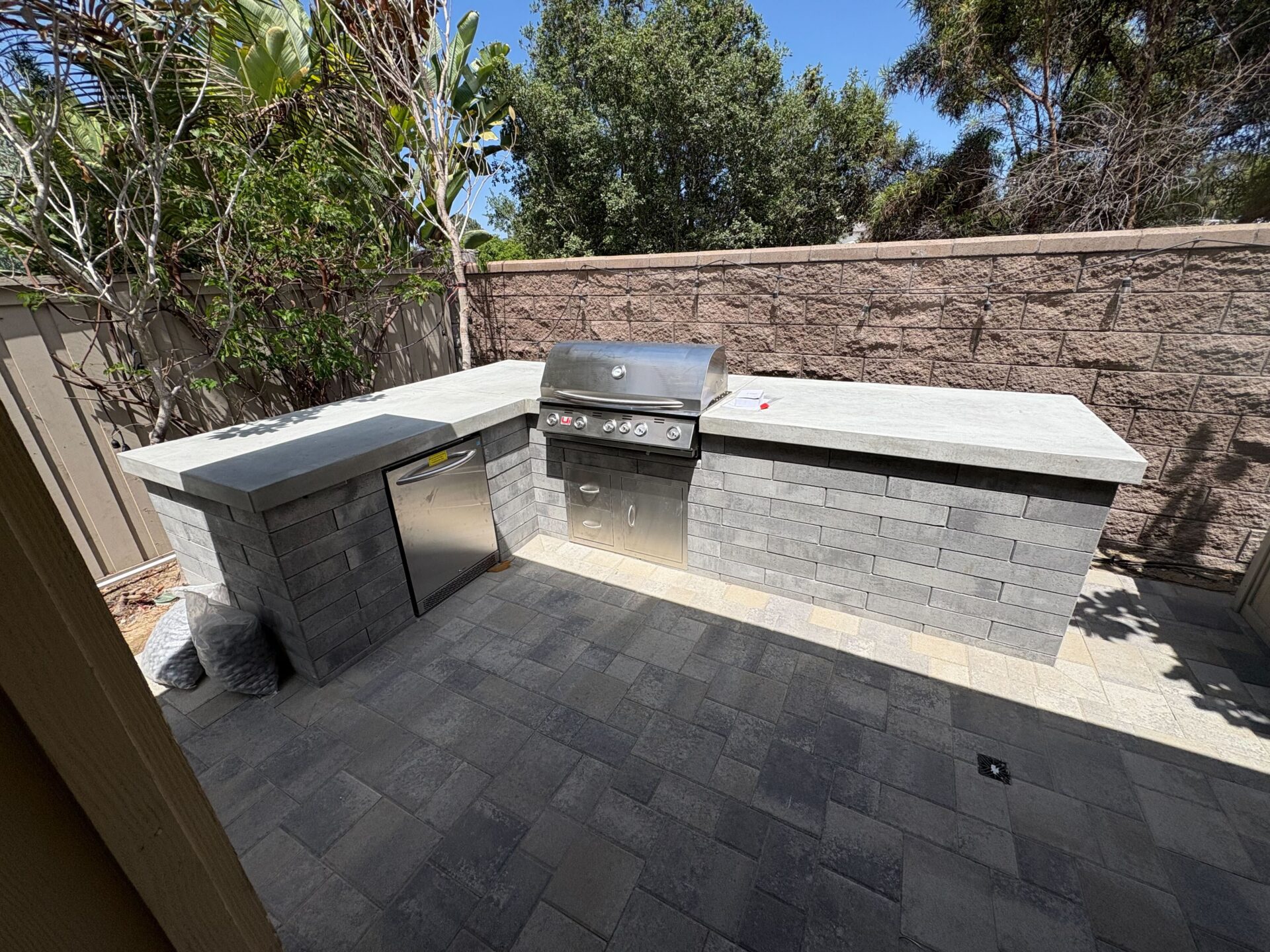 Finished outdoor kitchen with concrete counters, stainless grill, refrigerator, and paver patio.