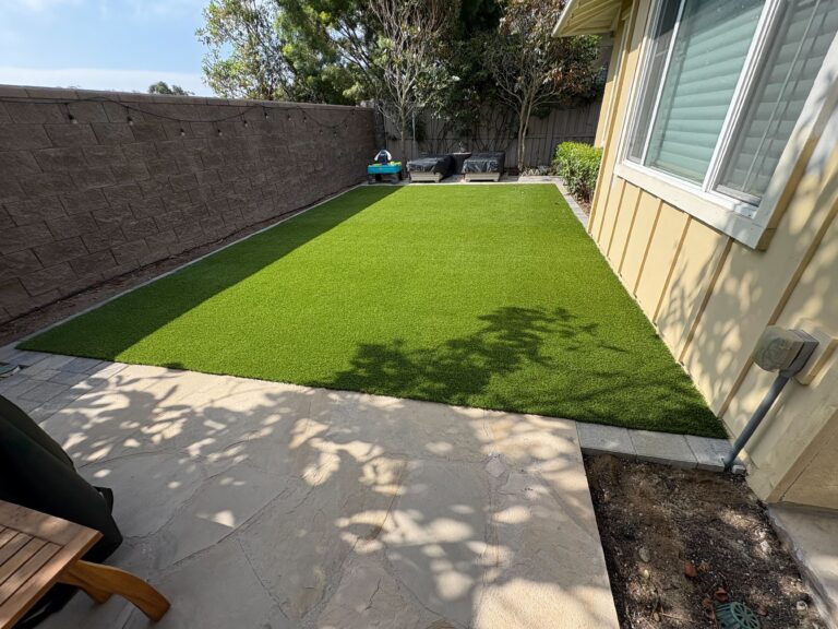 A finished residential backyard showing newly installed artificial turf bordered by a paved patio slab and a low...