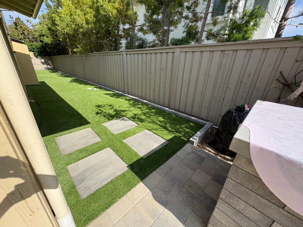 Finished narrow side yard showing installed artificial turf with rectangular concrete stepping pads set flush in the turf...