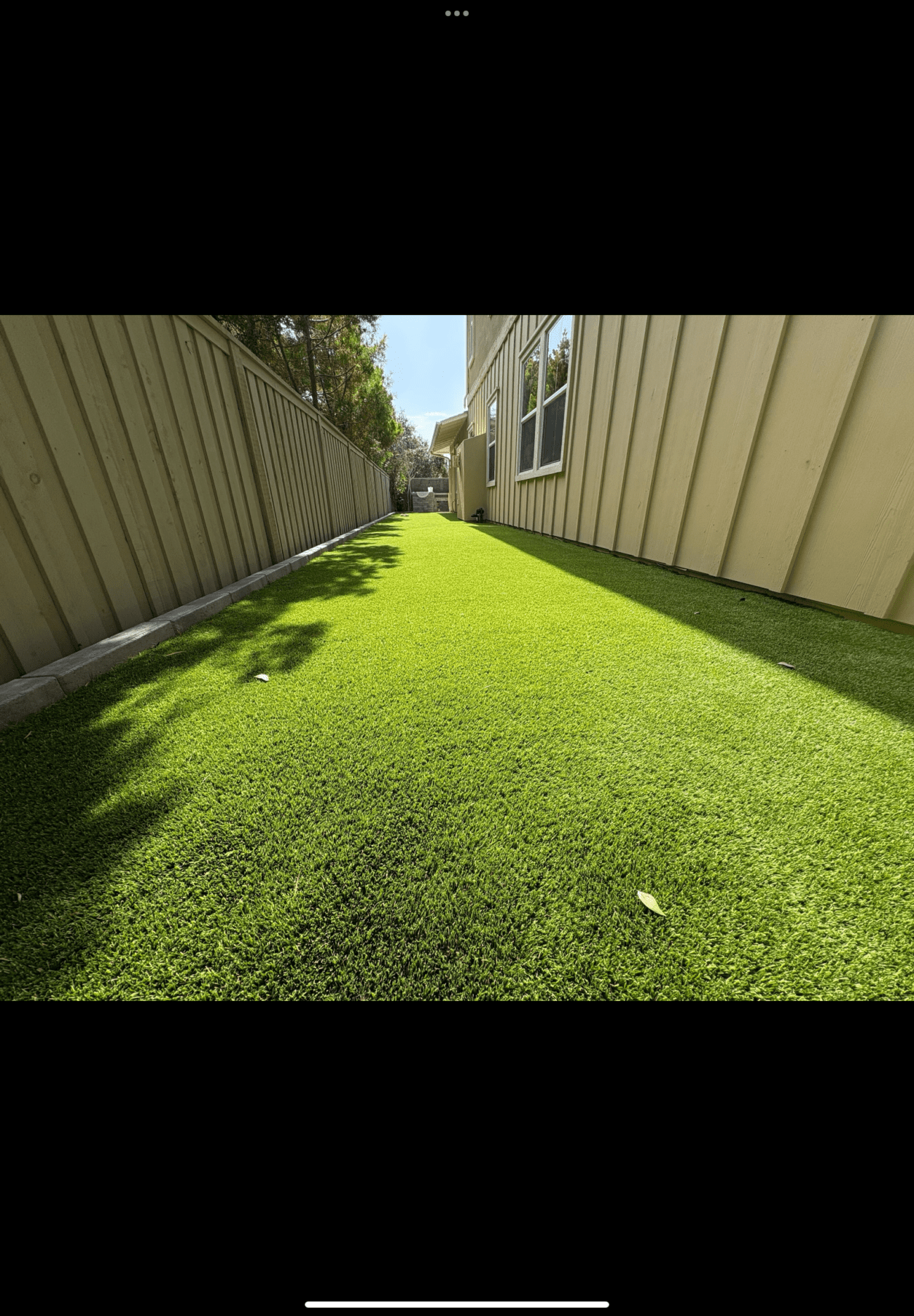 Completed artificial turf installation in a narrow side yard between a house and fence. The synthetic grass is uniformly...