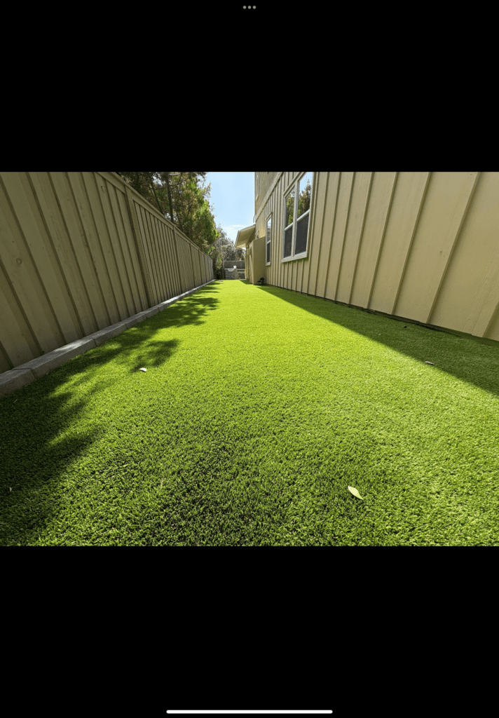 Completed artificial turf installation in a narrow side yard between a house and fence. The synthetic grass is uniformly...